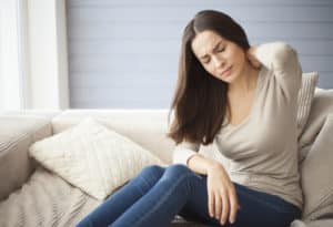 young brunette girl sitting on the couch at home with a headache and back pain.